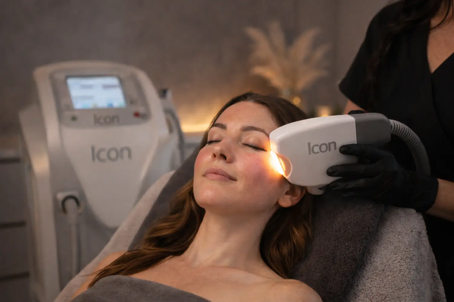 Practitioner holding a handheld device to a woman’s cheek while she reclines in a treatment chair.