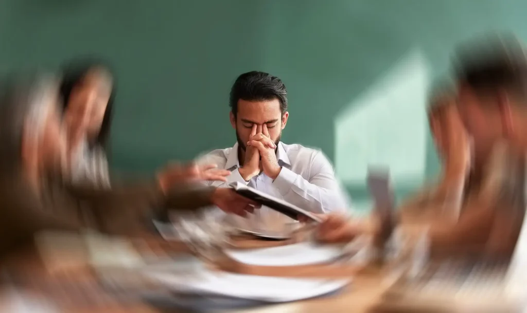 Man sits with eyes closed and hands clasped near his face while people around him appear blurred.